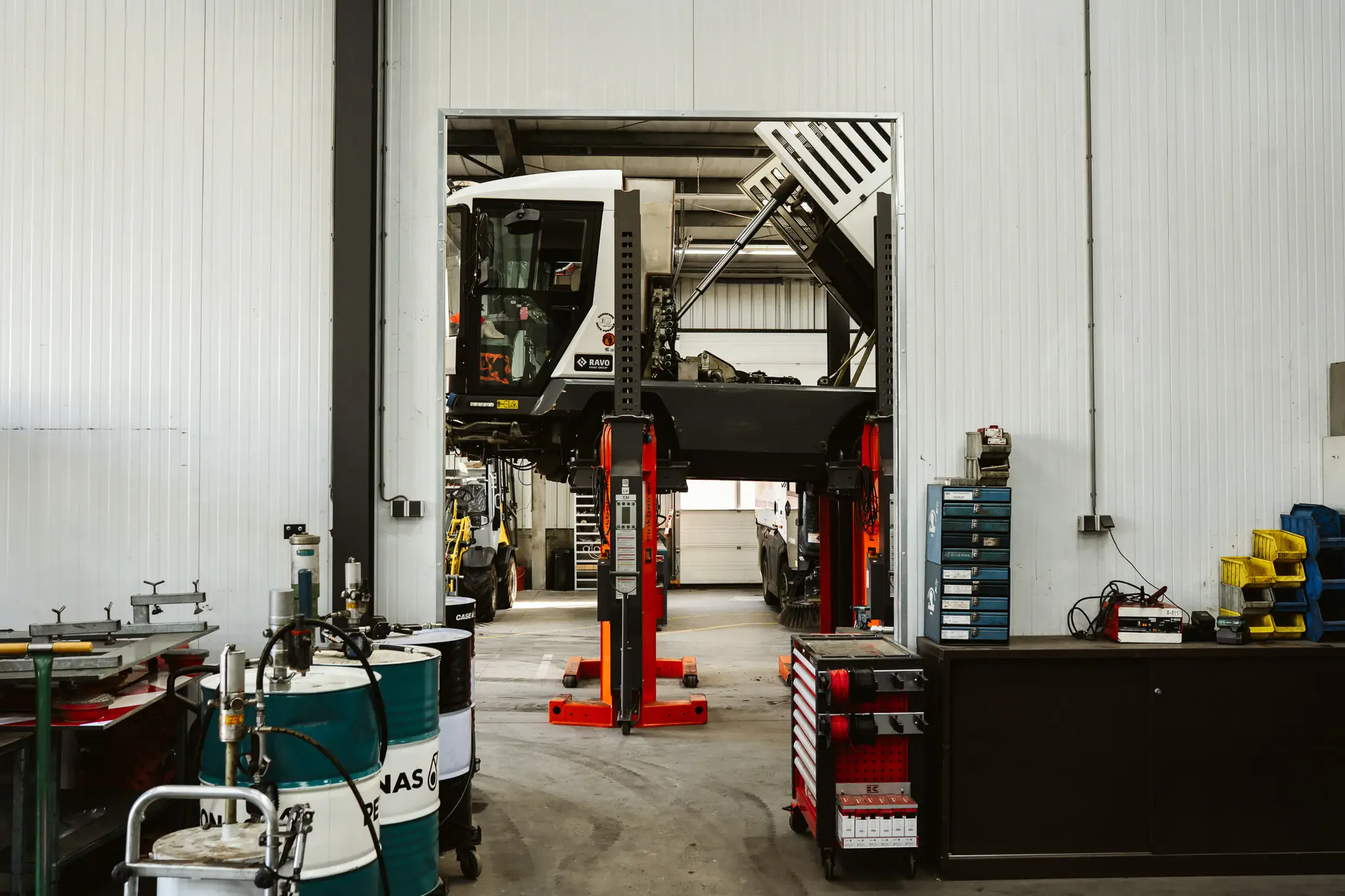 Doorkijk vanuit de werkplaats naar een veegmachine op hefkolommen met de opvangbak omhoog.
